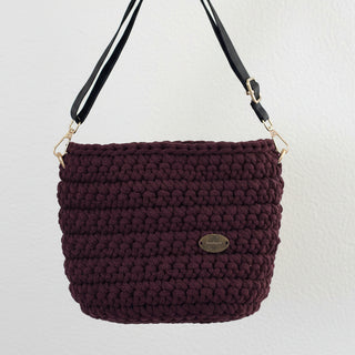 A handmade crochet crossbody bag in burgundy color with a black strap and a brand tag visible on the side.