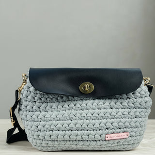 A gray crochet crossbody bag with a leather flap, displayed on a wooden surface.