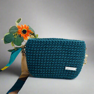 A green toned, crochet handbag with a textured pattern, displayed on a white surface with a decorative flower and a tag showing.