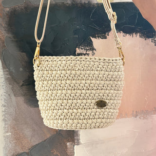 A small handmade crochet crossbody bag in shiny ivory color with a leather strap, displayed against an abstract painting background.