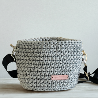 A handmade crochet crossbody bag in silver color with a subtle shimmer, featuring a black strap and a tag showing the brand name.