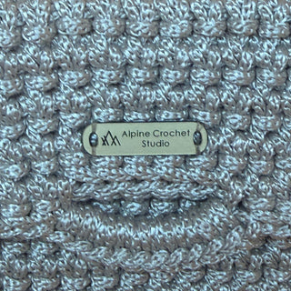 Close-up of a textured crocheted item in a heathered light gray and white yarn, featuring a metal brand plaque that reads 'Alpine-Crochet Studio'.