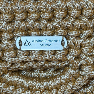 Close-up of a chunky crocheted textile in a warm tan and cream colorway, with a small white fabric label reading 'Alpine Crochet Studio' sewn into the crochet pattern.