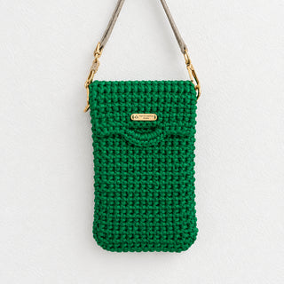 A green crocheted crossbody phone bag with a light gray strap and gold hardware, held by a hand against a plain white wall.