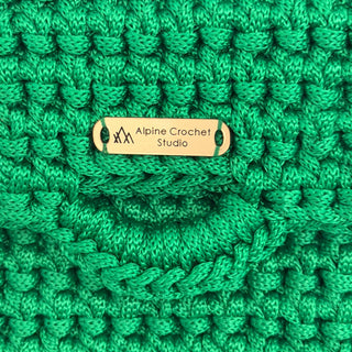 Close-up of a bright green crocheted textile with a small gold metal tag reading 'Alpine Crochet Studio'.
