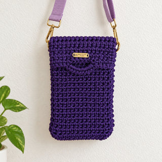 A purple crocheted phone pouch with a matching lavender strap and gold-tone hardware, hanging against a plain off-white wall.