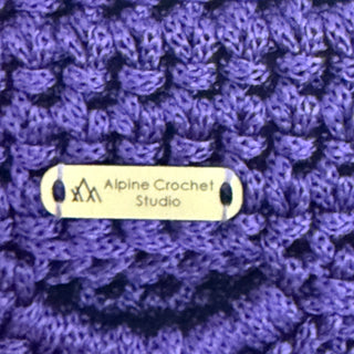 Close-up of a purple crocheted fabric with a small beige metal tag reading 'Alpine Crochet Studio' sewn into it.