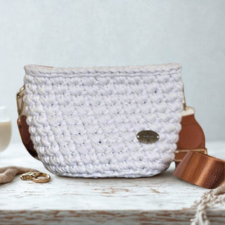 A white handmade crochet crossbody bag with a brown strap, displayed on a wooden surface. The bag has a solid color and a small brand tag attached to it.