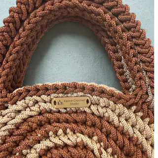 A close-up of a hand-crocheted bag with thick braided handles in a warm brown and cream colorway. A small wooden brand tag reading 'Alpine Crochet Studio' is sewn onto the front.