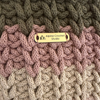 Close-up of a chunky knit fabric with three color sections: olive green, dusty pink, and light beige, featuring a small gold metal brand tag that reads 'Alpine Crochet Studio'.