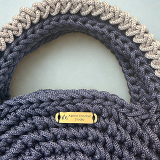 Close-up of a chunky crocheted handbag in dark gray with a contrasting light beige braided trim along the top edge and a small gold metal brand plaque reading 'Aline Crochet Studio'.