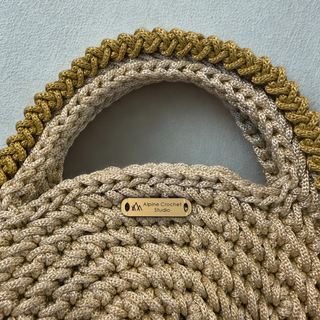 Close-up of a chunky crocheted handbag in beige with a wide braided handle in olive green and beige, featuring a small wooden brand plaque that reads 'Aspire Crochet Studio'.