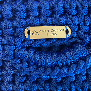 A close-up of a vibrant royal blue chunky crochet fabric with a small gold metal brand tag reading 'Alpine Crochet Studio' in the center.