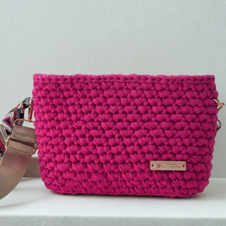 A handmade crochet crossbody bag in hot pink, made from cotton T-shirt yarn, displayed on a white shelf.